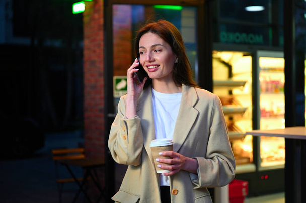 Günlük kıyafetler giymiş güzel bir kadın şehir ortamında bir fincan kahvenin tadını çıkarıyor. Telefonuyla konuşurken derin bir sohbete dalmış gibi görünüyor. Hem odaklanmış hem de memnun olduğunu gösteren bir ifadeyle.. - Fotoğraf, Görsel