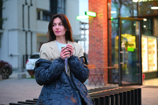 Krásná žena oblečená v neformálním oblečení sedí v městském prostoru a vychutnává si šálek kávy. Obklopuje ji městský ruch, ale zůstává klidná a vyrovnaná, když si vychutnává svůj drink.. - Fotografie, Obrázek