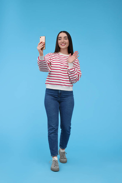 Smiling young woman taking selfie with smartphone on light blue background - Foto, afbeelding