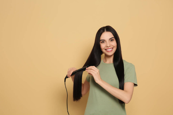 Beautiful happy woman using hair iron on beige background. Space for text - Zdjęcie, obraz