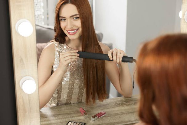 Beautiful woman using hair iron near mirror in room - Foto, afbeelding