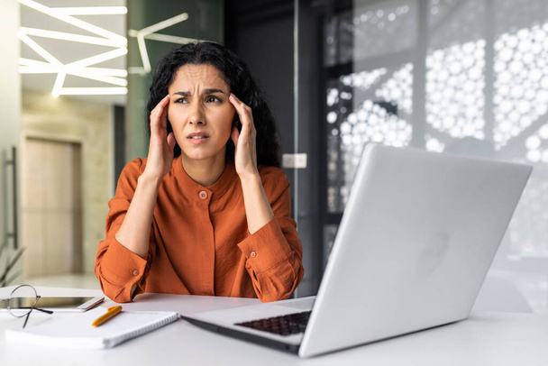 Beautiful woman at workplace overworked, tired hispanic woman at workplace with severe headache, businesswoman working inside office sitting with laptop, stressed and sad. - 写真・画像