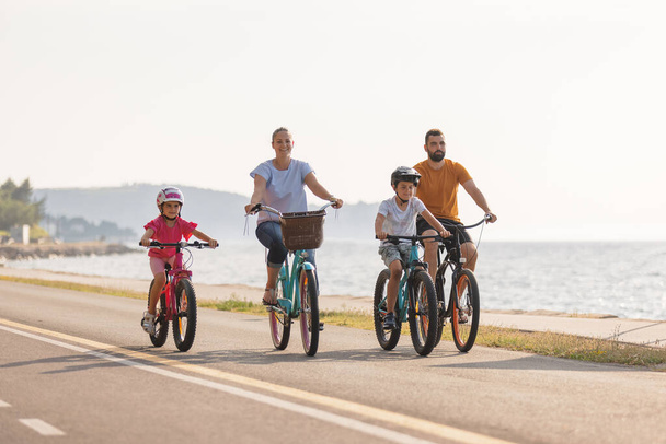 Two kids, with helmets on their heads, and smiling parents riding bikes on a family-friendly cycle route along a sea coastline, front view. - Fotó, kép