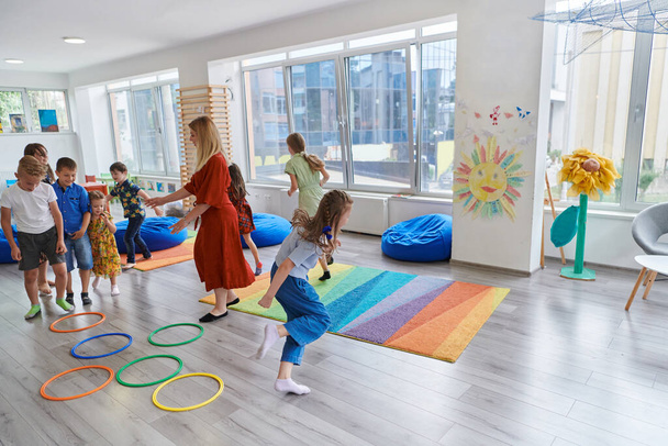 Small nursery school children with female teacher on floor indoors in classroom, doing exercise. Jumping over hula hoop circles track on the floor - Fotografie, Obrázek