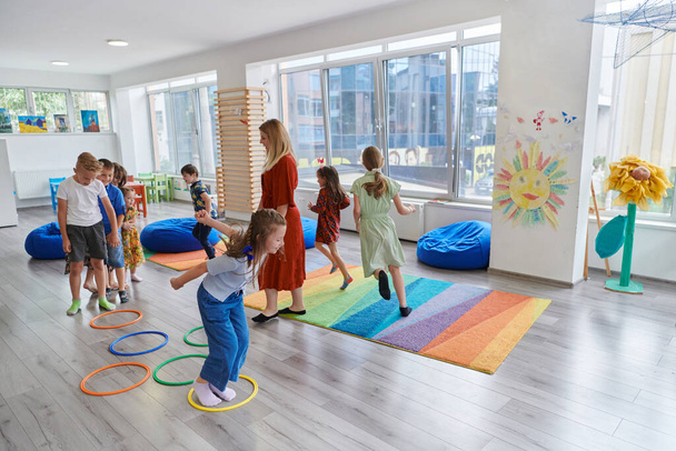 Small nursery school children with female teacher on floor indoors in classroom, doing exercise. Jumping over hula hoop circles track on the floor - Foto, imagen