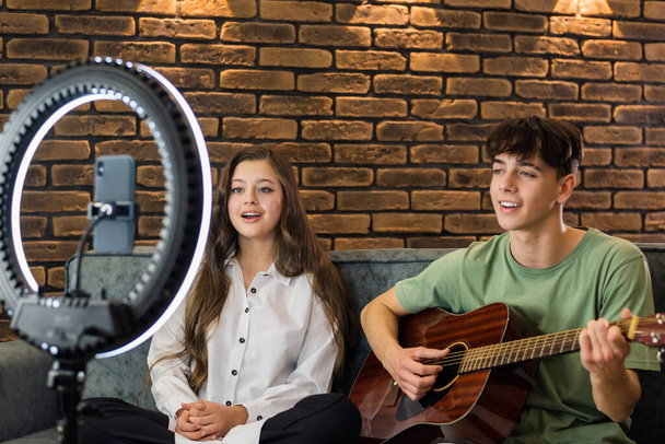 Young smiling man and woman playing on a guitar and singing, shooting themselves on smartphone on a tripod with ring light in a studio - Fotoğraf, Görsel