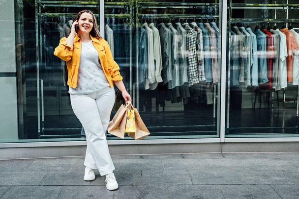 Plus size woman with shopping bags talking by cell phone on shop window background. Shopping woman talking on the smartphone. - Valokuva, kuva