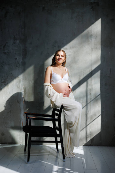 Egy fehér öltönyös, elegáns, fiatal terhes nő ül egy szürke hátterű széken. Terhes nő fehér fehérneműben. - Fotó, kép