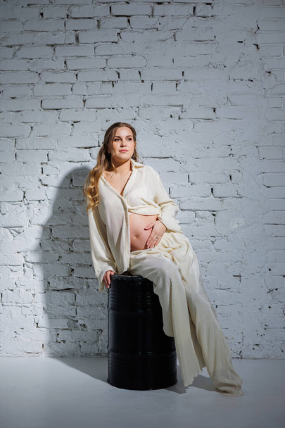 A stylish pregnant young woman in a white suit sits against a white brick wall and touches her belly with a gentle smile. Awaiting birth. Care and motherhood. Love and tenderness. - Photo, Image