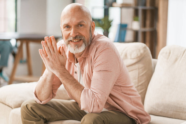 Smiling, happy senior man sitting on a sofa in the comfort of his home. With his hands held together, he radiates a sense of contentment and relaxation. . High quality photo - Fotoğraf, Görsel