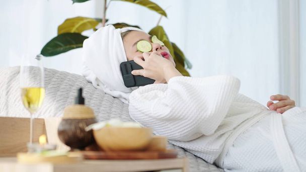 Жінка, що лежить на спа-ліжку, відчуває розслаблення зі скибочками огірків на очах і розмовляє по смартфонах у спа-кімнаті. Концепція спа-ароматерапії
 - Фото, зображення