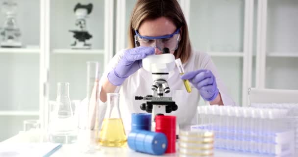 La mujer química ajusta el microscopio sosteniendo el tubo con líquido amarillo en la mano. Mujer experimenta paciente muestra de orina sentada en la mesa en la oficina cámara lenta - Imágenes, Vídeo