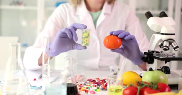 Nainen kemisti kumihanskat pitää pakkaus pillereitä ja tuoretta orgaanista mandariinia. Vitamiinien hyödyllisyyden käsite tableteissa ja sitrushedelmissä hidastettuna - Materiaali, video