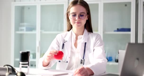 Positiivinen nainen lääkäri osoittaa punainen sydän lelu kädessä istuu klinikan toimistossa. Nainen brunette henkilö stetoskooppi hieman hymyillen hidastettuna - Materiaali, video