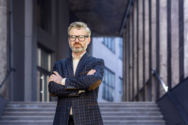 Portrait of mature successful boss, gray haired businessman looking at camera with crossed arms, man outside office building in business suit and glasses, satisfied and focused banker investor - Foto, Bild