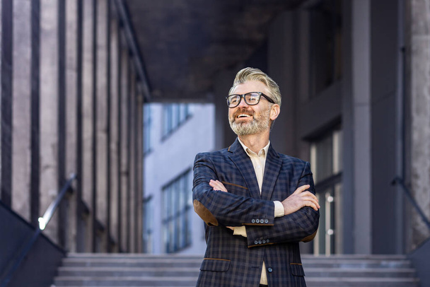 Portrait of mature adult gray-haired businessman, man with crossed arms outside office building smiling and looking away, boss in business suit successful entrepreneur. - Valokuva, kuva