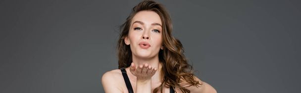 Ładny młody model z naturalnym makijażem i brunetka włosy dmuchanie pocałunek powietrza podczas flirtu i patrząc w obiektyw stojąc odizolowany na szary, baner  - Zdjęcie, obraz