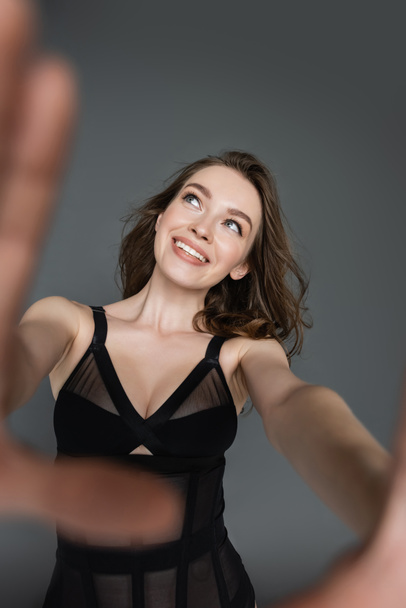 Smiling and fashionable young brunette woman with natural makeup in sexy black bodysuit looking away while posing and standing isolated on grey, blurred hands  - Фото, зображення