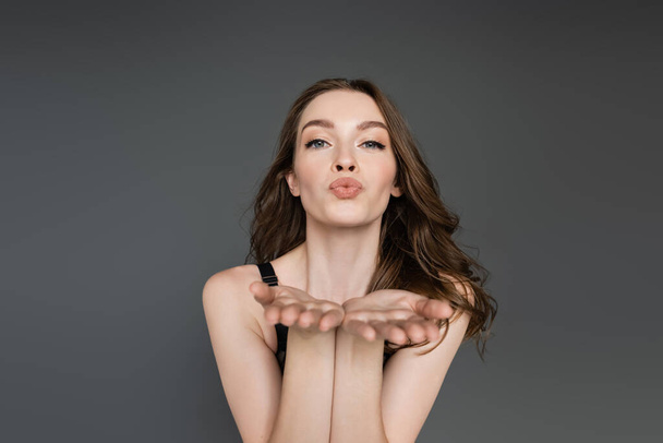 Pretty and young brunette model with hairstyle and natural makeup looking at camera while blowing air kiss while standing and posing isolated on grey,  flirt, chic  - Photo, Image