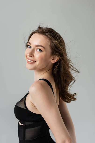 Portrait of positive and pretty young brunette woman with brunette hair and natural makeup looking at camera while posing in black bodysuit isolated on grey   - Foto, Imagem