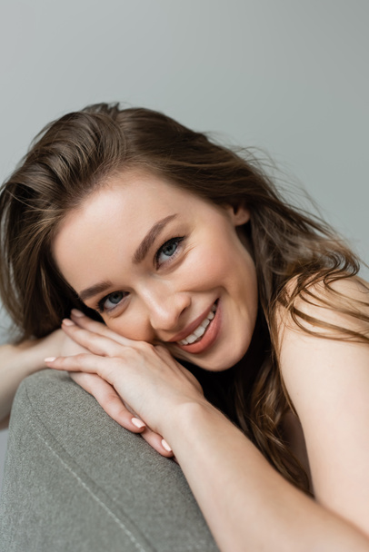 Portrait of smiling and pretty young woman with natural makeup and brunette hair looking at camera while relaxing on comfortable grey armchair isolated on grey   - Foto, Imagem