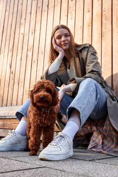 Ένα νεαρό χαμογελαστό ξανθό κορίτσι με τζιν και ένα μπεζ αδιάβροχο κάθεται και παίζει με το σκυλάκι της ενώ περπατάει στο δρόμο στο φόντο ενός ξύλινου τοίχου. Περπατήστε και να χαλαρώσετε με το κατοικίδιο ζώο σας - Φωτογραφία, εικόνα