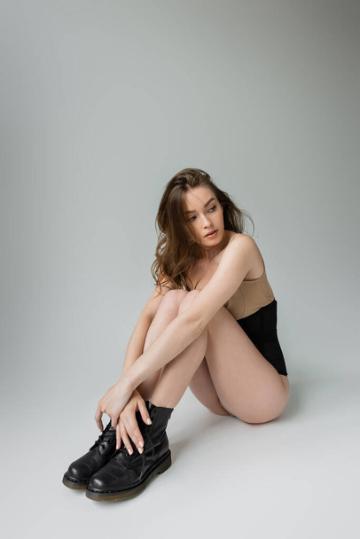 Full length of sexy and fashionable young woman in black boots, beige bodysuit and corset looking away while sitting and posing on grey background  - 写真・画像