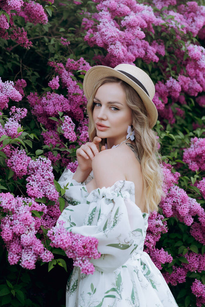Beautiful, stylish, cute blonde in a white dress with a green pattern as a model for a photo shoot in lilac. They made luxurious portraits of her in the green garden of the park. - Valokuva, kuva