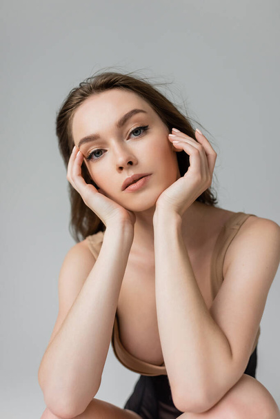 Portrait of young and seductive woman with brunette hair and natural makeup wearing bodysuit while touching face and looking at camera isolated on grey  - Foto, immagini