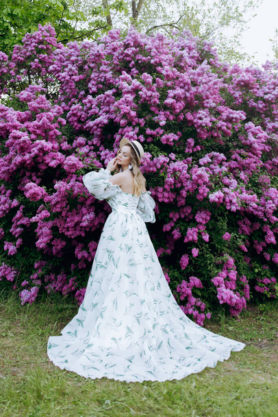Schöne, stylische, süße Blondine in einem weißen Kleid mit grünem Muster als Modell für ein Fotoshooting in Flieder. Sie machten luxuriöse Porträts von ihr im grünen Garten des Parks. - Foto, Bild