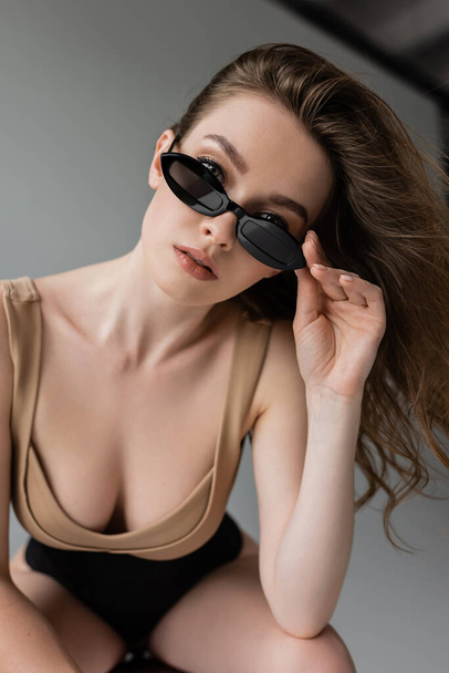 Portrait of stylish and sexy brunette woman with natural makeup touching sunglasses and looking at camera while posing in beige bodysuit and corset on grey background  - 写真・画像