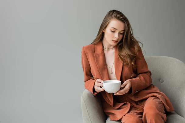 Módní mladá žena v terakotové bundě a kalhoty nosí náhrdelníky a drží šálek cappuccino, zatímco sedí na pohodlném křesle izolované na šedé   - Fotografie, Obrázek