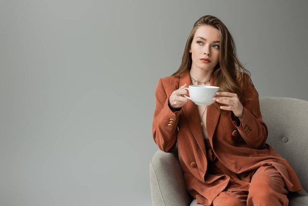 Trendy young and brunette woman in terracotta suit holding cup of cappuccino and looking away while sitting on modern armchair isolated on grey with copy space - Photo, image
