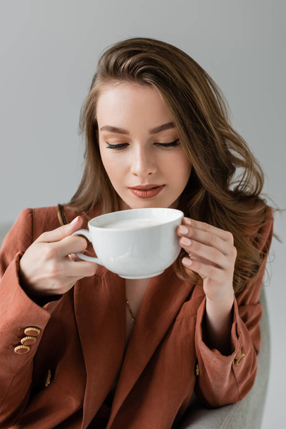 brunette young woman with long hair wearing trendy suit with blazer and holding cup of coffee while sitting in comfortable armchair on grey background in studio, work-life balance, terracotta  - Foto, imagen