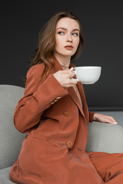 young woman with long hair wearing brown and trendy suit with blazer and pants and holding cup of coffee while sitting in comfortable armchair on grey background, looking away, work-life balance - Photo, Image