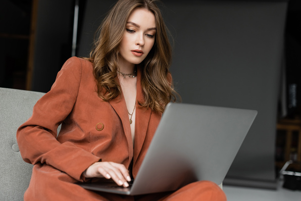 barna fiatal nő hosszú haj és nyaklánc visel terrakotta elegáns öltöny blézer és nadrág segítségével laptop, miközben ül kényelmes karosszék szürke háttér, szabadúszó, távoli munka  - Fotó, kép