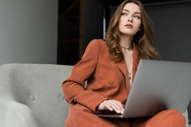 pensive young woman with long hair and necklace wearing trendy suit with blazer and pants and using laptop while sitting in comfortable armchair on grey background, freelancer, remote work  - Photo, Image