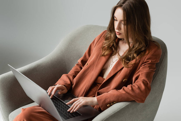 brunette young woman with long hair and necklace wearing terracotta trendy suit with blazer and pants and typing on laptop while sitting in comfortable armchair on grey background, remote work - Photo, Image