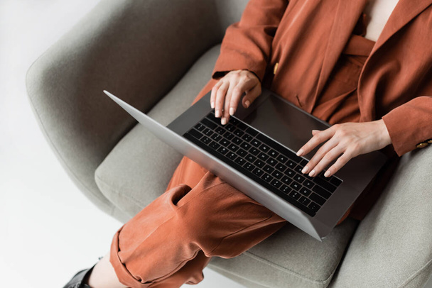 Draufsicht der jungen Frau im trendigen Anzug mit Blazer und Hose mit Laptop im bequemen Sessel auf grauem Hintergrund sitzend, Freiberuflerin, Fernarbeit, Schnappschuss  - Foto, Bild
