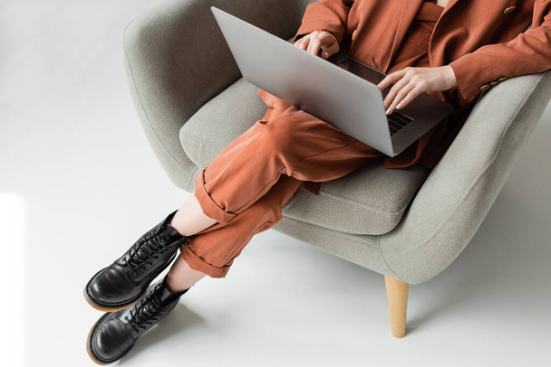 top view of young woman wearing terracotta trendy suit with blazer and pants with boots using laptop while sitting in comfortable armchair on grey background, freelancer, remote work, cropped shot - Photo, Image
