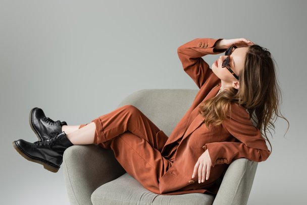 mujer joven y elegante con pelo largo que lleva traje de terracota con chaqueta, pantalones y botas negras posando en gafas de sol de moda mientras está sentado en un sillón sobre fondo gris, modelo de moda  - Foto, imagen