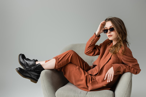 κομψή νεαρή γυναίκα με μακριά μαλλιά φορώντας κοστούμι τερακότα με σακάκι, παντελόνι και μαύρες μπότες ποζάρουν σε μοντέρνα γυαλιά ηλίου, ενώ κάθεται στην πολυθρόνα σε γκρι φόντο, μοντέρνο μοντέλο, κοιτάζοντας κάμερα - Φωτογραφία, εικόνα