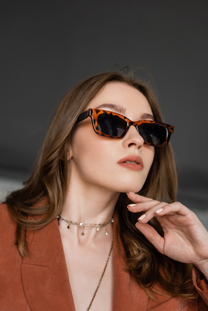 portrait of stylish young woman with long hair wearing suit with terracotta blazer and posing in trendy sunglasses while looking away near blurred armchair on grey background, fashionable model  - Foto, Imagem