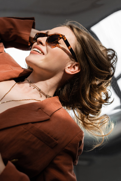 top view of happy young woman with long hair and natural makeup wearing terracotta and trendy suit with blazer and golden necklace while posing in sunglasses on grey background, beautiful model  - Foto, imagen