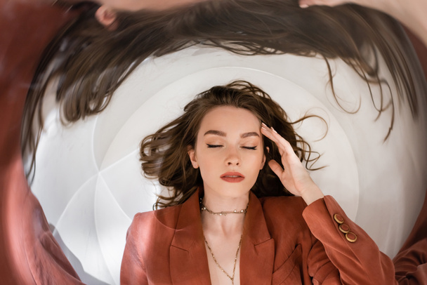 circular shot, top view of young woman with brunette hair wearing blazer and golden necklace while lying and touching face on grey background, blurred, wide-angle, distorted view, closed eyes - Photo, Image