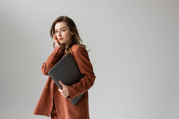 pretty young woman with brunette hair and necklace wearing terracotta and trendy suit with blazer, looking at camera and holding laptop while standing on grey background, freelancer, remote work  - Photo, Image