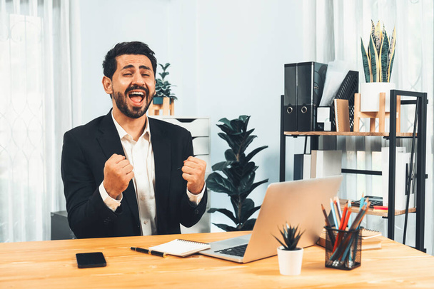 Возбужденный и счастливый бизнесмен, одетый в черный официальный костюм, поднимает руку в праздничном жесте за рабочим столом, после успешного продвижения по службе, энергии и радости в качестве бизнес-победителя. Пылкий - Фото, изображение