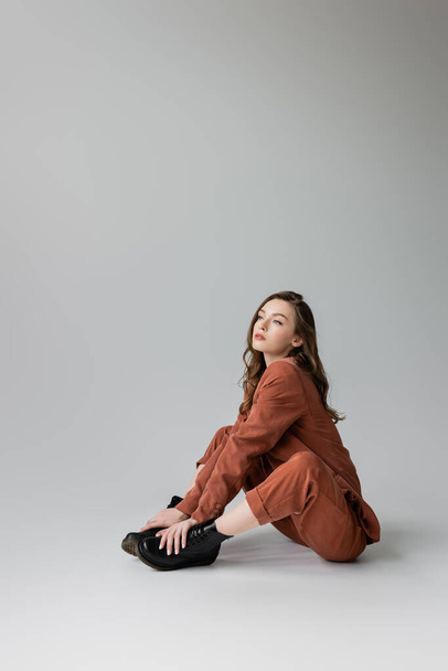 full length of dreamy model with brunette and wavy hair sitting in trendy and oversize terracotta suit with blazer, pants and black boots, looking away on grey background, young woman, stylish pose - Fotoğraf, Görsel