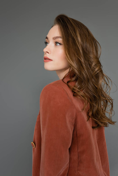 portrait of sophisticated young woman with brunette hair posing in brown and trendy suit with blazer while looking away on grey background in studio, pretty model, curls, trendy hairstyle  - Φωτογραφία, εικόνα