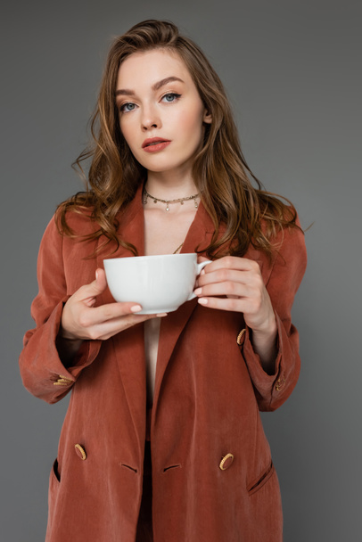 jeune femme sophistiquée aux cheveux bruns portant un costume marron et tendance avec un blazer et tenant une tasse de café tout en regardant la caméra sur fond gris, équilibre travail-vie privée  - Photo, image